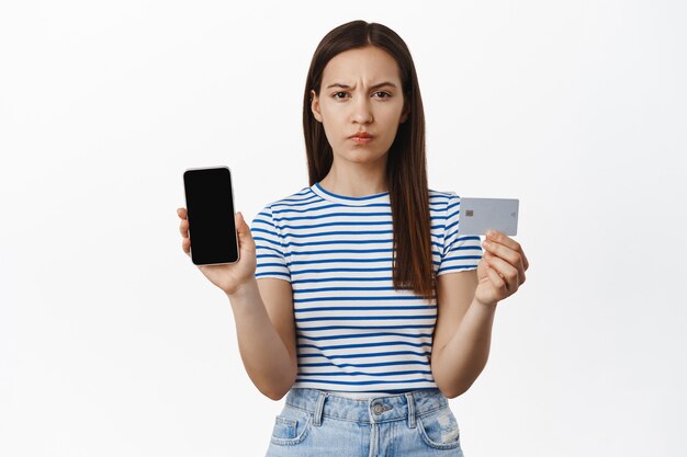 화난 여성은 눈살을 찌푸리고 신용 카드와 휴대폰 빈 화면, 은행 계좌 또는 스마트폰 앱 인터페이스를 보여주고 눈썹을 찡그리며 불쾌해하고 흰 벽을 보여줍니다.