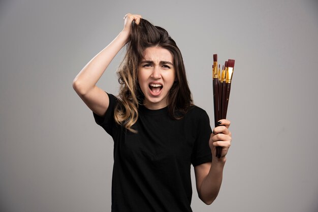 Angry woman holding brushes touching her hair  on gray wall.