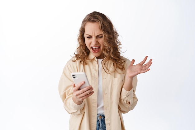 화난 여성이 스마트폰 화면을 보고 흰색 배경 위에 서 있는 휴대전화에 화난 표정으로 불평