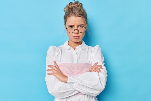 Angry woman mad as evil. Unhappy offended female model has bothered hateful expression crosses arms against chest stands in defensive pose wears spectacles and white shirt isolated on blue wall