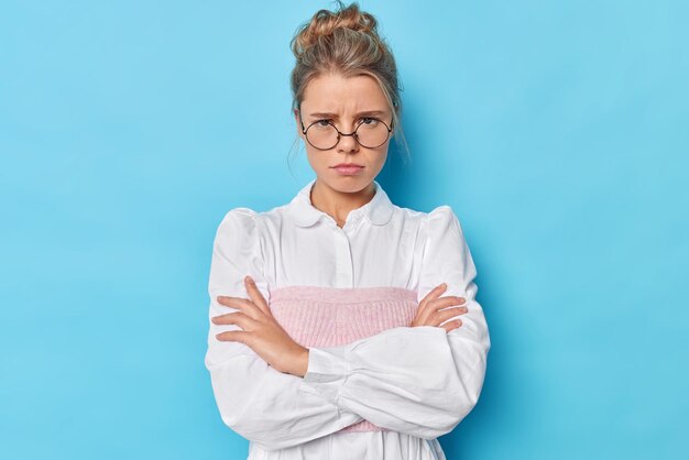 Angry woman mad as evil. Unhappy offended female model has bothered hateful expression crosses arms against chest stands in defensive pose wears spectacles and white shirt isolated on blue wall