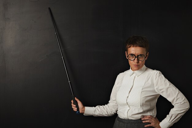 Angry woman teacher and shows on black chalk board behind her with folding pointer