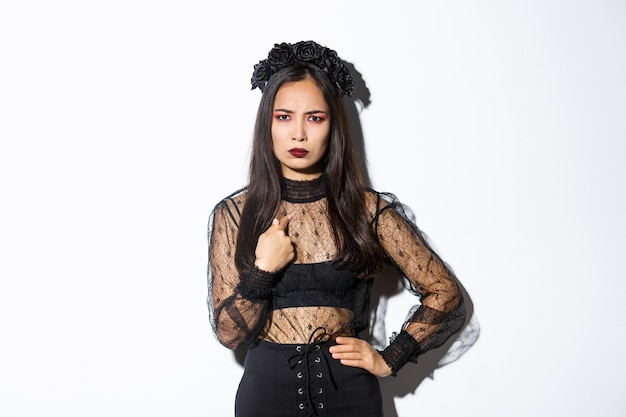 Angry woman in witch costume pointing at herself, frowning upset, standing frustrated in halloween costume, standing over white background.