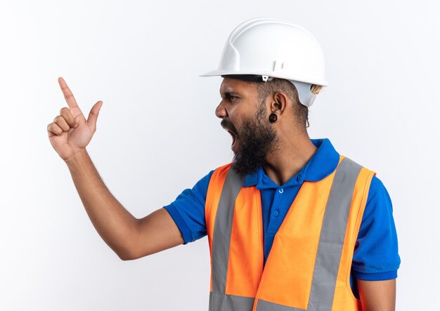 Angry young afro-american builder man in uniform with safety helmet yelling at someone looking at side isolated on white background with copy space