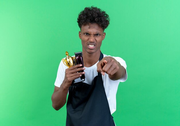 Angry young afro-american male barber wearing uniform holding cup winner and showing you gesture isolated on green background