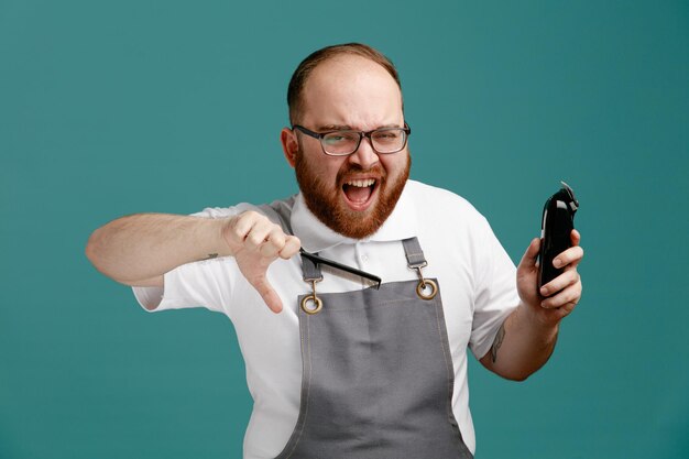 Angry young barber wearing uniform and glasses holding teaser comb and hair trimmer looking at camera showing thumb down isolated on blue background