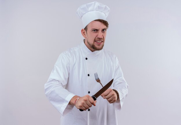 An angry young bearded chef man wearing white cooker uniform and hat holding knife and fork in x sign while looking on a white wall
