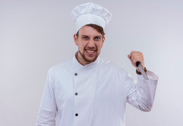 白い壁を見ながら白い炊飯器の制服と帽子保持ナイフを身に着けている怒っている若いひげを生やしたシェフの男
