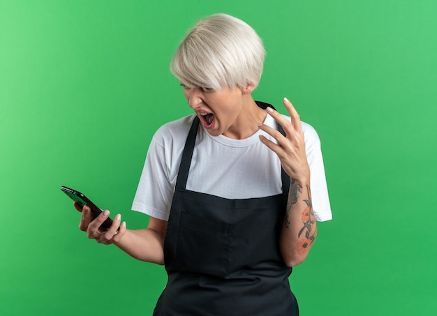 Giovane e bella barbiere femminile arrabbiata in uniforme che tiene e guarda il telefono isolato sul muro verde