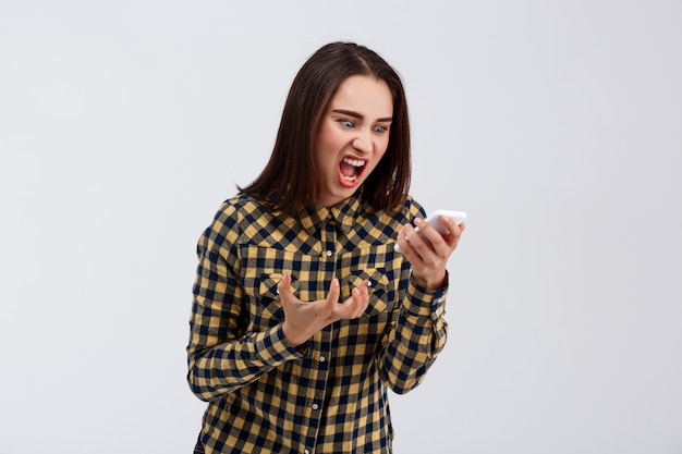 Angry young beautiful girl dressed in plaid shirt screaming at phone over white wall.