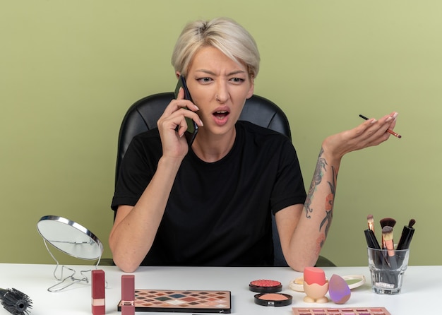 Angry young beautiful girl sits at table with makeup tools speaks on phone holding makeup brush isolated on olive green wall