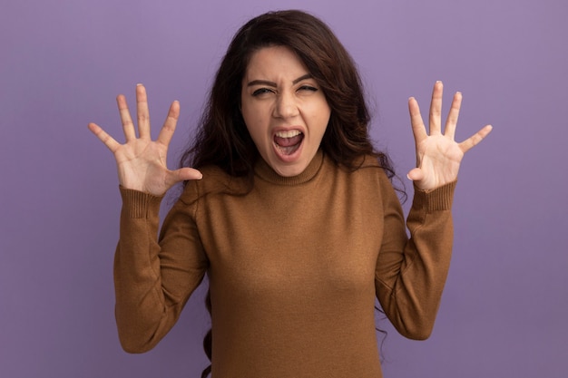 Free photo angry young beautiful girl wearing brown turtleneck sweater spreading hands isolated on purple wall