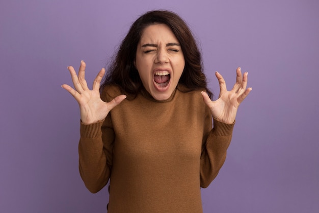 Foto gratuita giovane bella ragazza arrabbiata che porta maglione dolcevita marrone che diffonde le mani isolate sulla parete viola