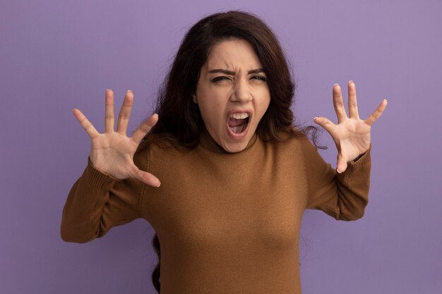 Angry young beautiful girl wearing brown turtleneck sweater spreading hands isolated on purple wall