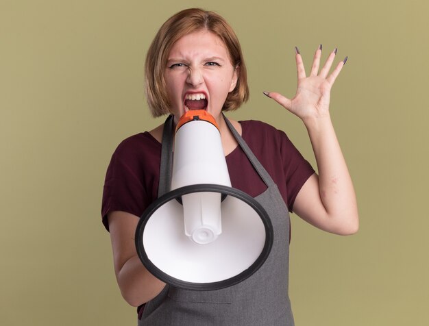Angry young beautiful woman hairdresser in apron shouting to megaphone with arm raised standing over green wall