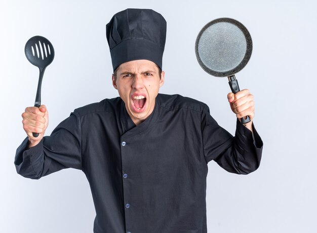 Angry young blonde male cook in chef uniform and cap looking at camera showing spatula and frying pan screaming isolated on white wall