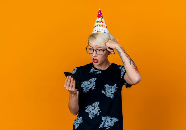 Сердитая молодая блондинка вечеринка в очках и кепке дня рождения держит и смотрит на мобильный телефон, касаясь головы, изолированной на оранжевой стене
