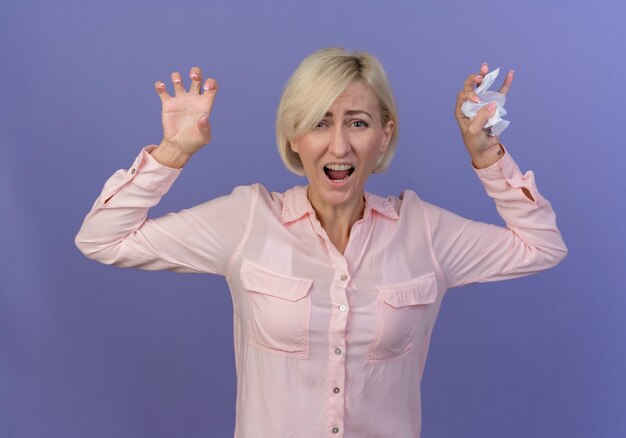 Angry young blonde slavic woman raising hands with crushed paper in hand isolated on purple background