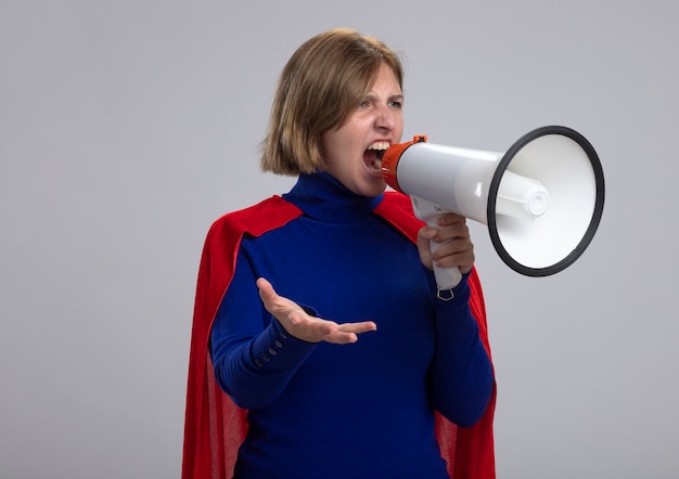 Angry young blonde superhero girl in red cape shouting in loud speaker looking straight showing empty hand isolated on white wall with copy space