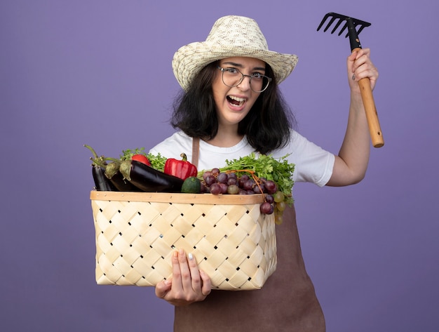 Angry young brunette female gardener in optical glasses and in uniform wearing gardening hat holds vegetable basket and rake isolated on purple wall