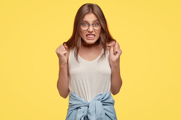 Angry young brunette with glasses posing against the yellow wall