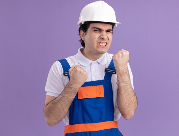 Angry young builder man in construction uniform and safety helmet clenching fists annoyed and irritated standing over purple wall