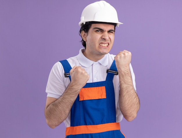 Злой молодой строитель в строительной форме и сжимая кулаки в защитном шлеме раздраженно и раздраженно стоит над фиолетовой стеной