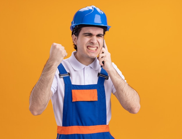 オレンジ色の壁の上に立っている携帯電話で話している建設制服と安全ヘルメットの怒っている若いビルダーの男