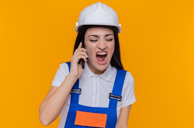 Angry young builder woman in construction uniform and safety helmet shouting going wild while talking on mobile phone standing over orange wall