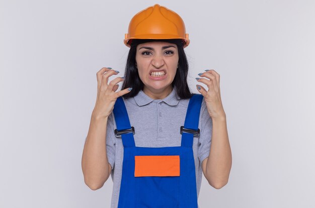 Angry young builder woman in construction uniform and safety helmet yelling with arms raised going wild