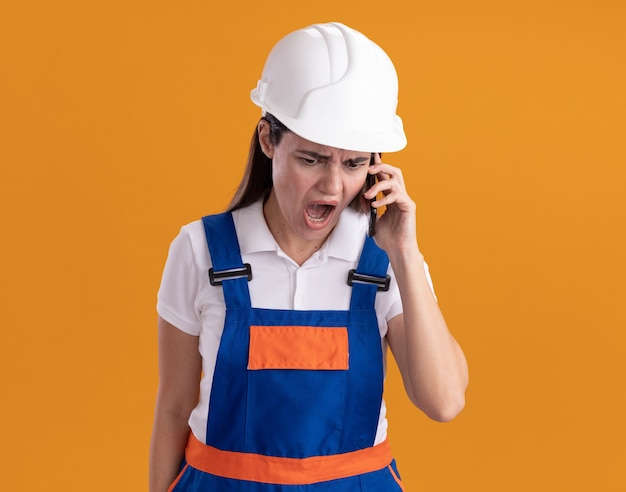 Angry young builder woman in uniform speaks on phone isolated on orange wall