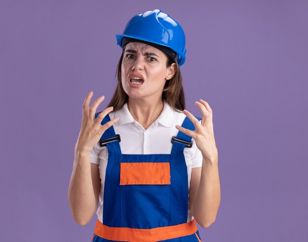 Angry young builder women in uniform holding hands around face isolated on purple wall