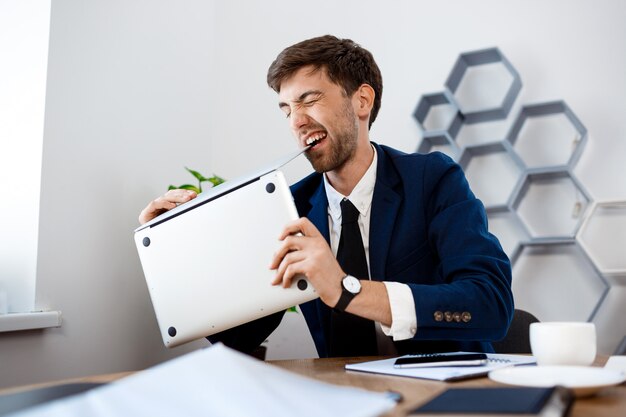 Angry young businessman gnawing laptop, office background.