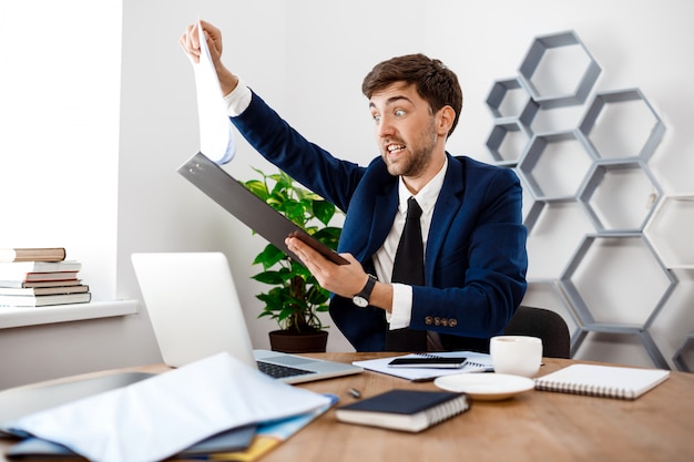 Angry young businessman sitting at workplace, office background.