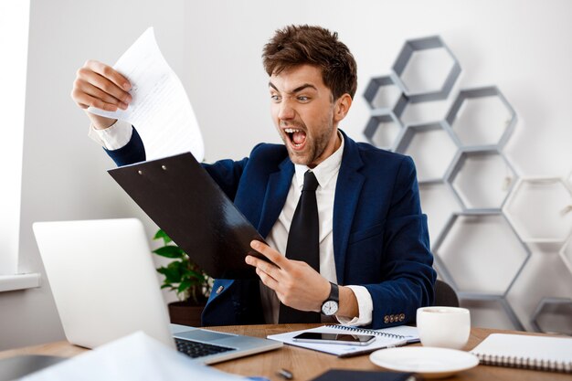 Angry young businessman sitting at workplace, office background.