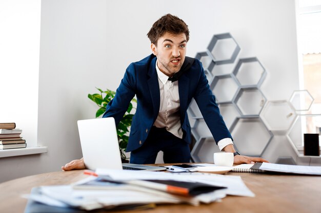 Angry young businessman standing at workplace, office background.