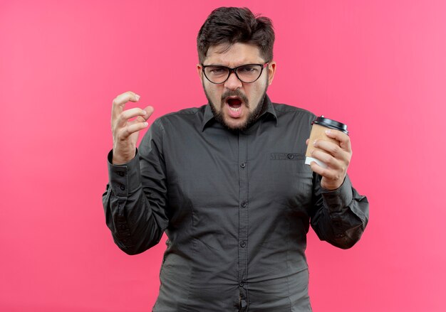 Angry young businessman wearing glasses holding cup of coffee isolated on pink wall