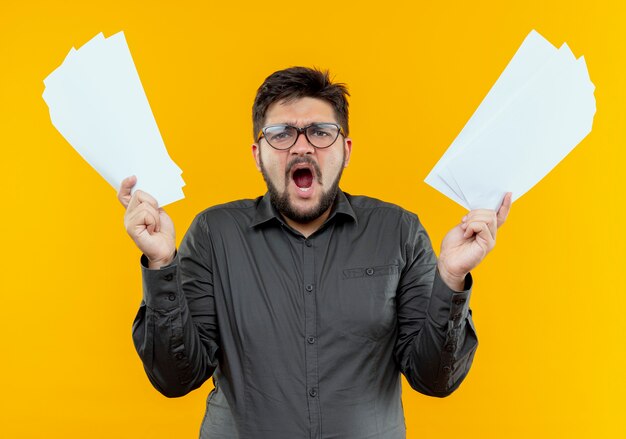 angry young businessman wearing glasses raising papers isolated on yellow