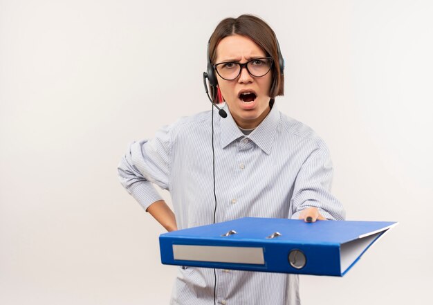 Angry young call center girl wearing glasses and headset stretching out clipboard putting hand on waist isolated on white  with copy space