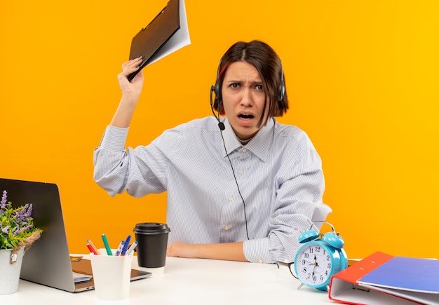 Angry young call center girl wearing headset sitting at desk raising up clipboard isolated on orange 