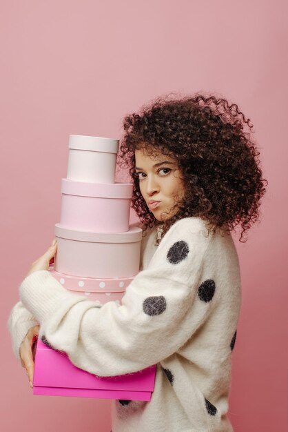 Angry young caucasian girl greedily holds gift boxes in her hands while standing sideways on pink background Beauty has curly black hair and white sweater Leisure relaxation and lifestyle concept