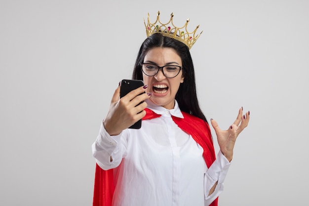 眼鏡と王冠を身に着けている怒っている若い白人のスーパーヒーローの女の子は、コピースペースで白い背景で隔離の空気中に手を保持している携帯電話を保持し、見ています