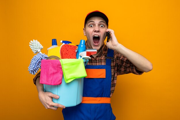 Angry young cleaning guy wearing uniform and cap holding bucket of cleaning tools speaks on phone 