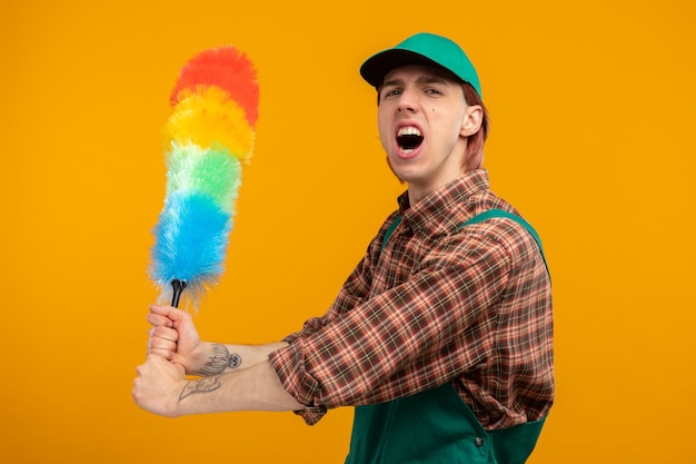 Angry young cleaning man in plaid shirt jumpsuit and cap swinging colorful duster shouting with aggressive expression standing on orange