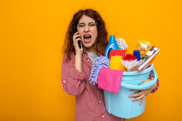 Angry young cleaning woman holding bucket of cleaning tools and speaks on phone 