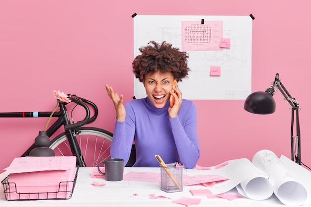 Angry young curly Afro American woman exclaims loudly has telephone conversation raises hand and screams from anger poses at coworking space being irritatated to make mistake in building sketch