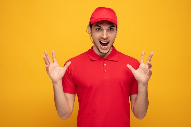 angry young delivery man wearing uniform and cap looking at camera showing empty hands screaming isolated on yellow background