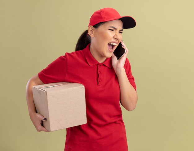 Сердитая молодая женщина-доставщик в красной форме и кепке держит картонную коробку и кричит во время разговора по мобильному телефону, стоя над зеленой стеной