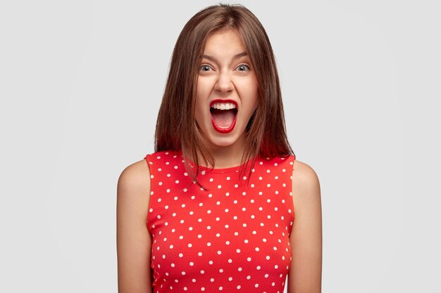 Angry young European female shouts with annoyance, has red lips, being annoyed with someone, dressed in fashionable dress, isolated over white wall. People and negative emotions concept