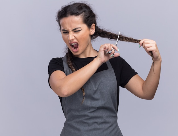 Angry young female barber in uniform cutting hair isolated on white wall